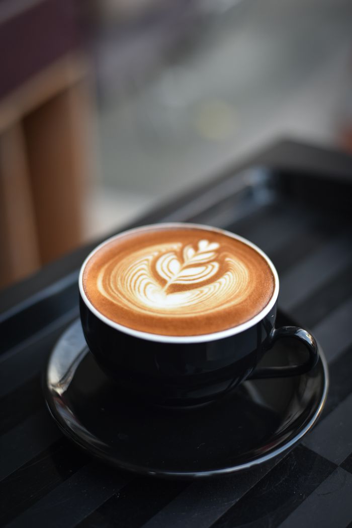 gallery-5 A beautifully crafted cappuccino with intricate latte art in a black ceramic mug on a dark surface.