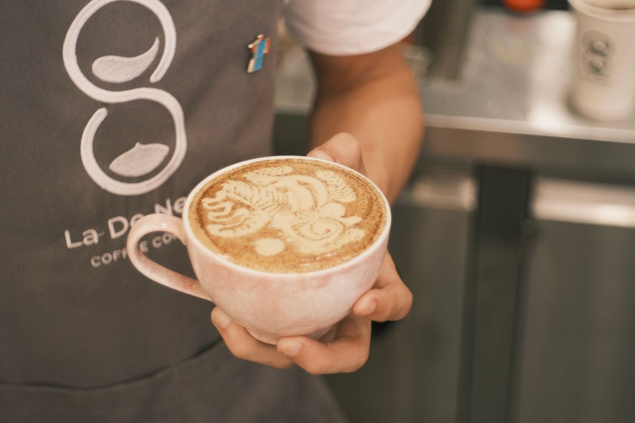hero-img-02 Close-up of a barista holding a cappuccino with intricate latte art, indoors in a coffee shop.