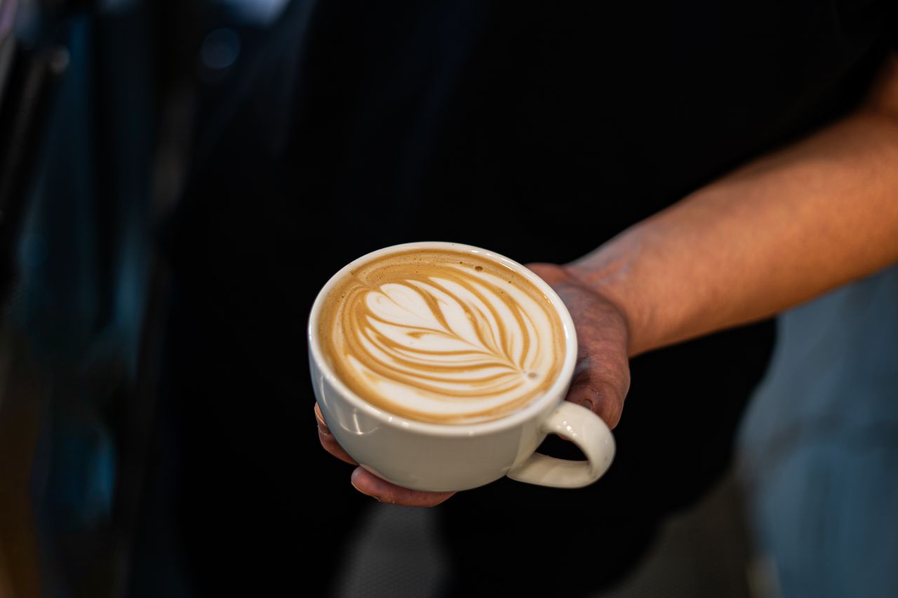 about-02 Close-up of a barista's hand holding a latte featuring intricate latte art.