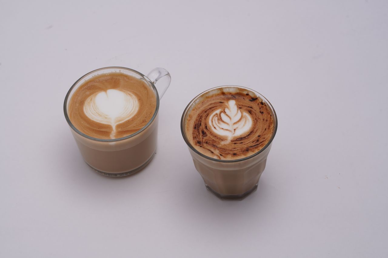 about-01 Two glass cups of latte with intricate latte art on white background.